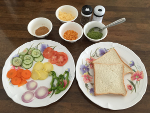 Preparation for layering ultimate Bombay vegetable sandwich, with cut vegetables, green sandwich chutney, butter, chaat masala, sev, cheeses and bread slice.